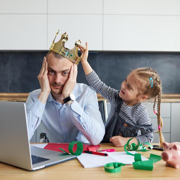 Tired Father Working from home on laptop during quarantine. Little child girl puts a toy crown on dads headon the kitchen office